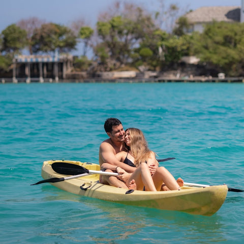 Couple in a yellow kayak on clear blue water with trees and a dock in the background.